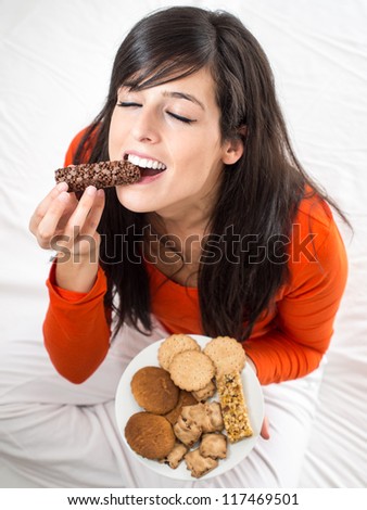 Woman eating chocolate bar. Hungry girl biting delicious  snack sitting on white bed indoor. Breaking diet concept.