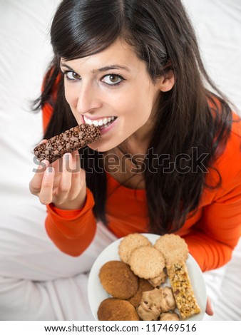Guilty happy woman eating chocolate bar. Beautiful brunette woman tasting  delicious crunchy snack sitting on white bed indoor.