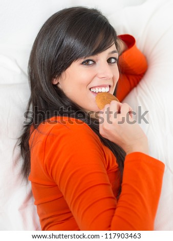 Beautiful young brunette woman eating biscuit and smiling lying down on white bed. Morning scene with casual playful cheerful hispanic girl.