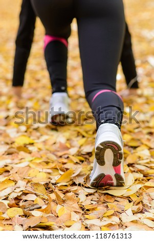 Sportswoman ready to run with powerful pose. Leg and foot in foreground. Wearing leggings and white trainers. Golden leaves on ground.
