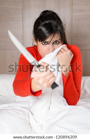 Frightened woman covering with the sheet on the bed and holding a knife in her hand. Brunette model looking scared and threatening in her room.