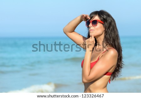 Worried woman on the beach watching with blue sea in the background. Beautiful brunette caucasian model with red sunglasses looking for someone.