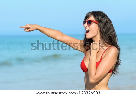 Beautiful brunette woman on the beach pointing happy to someone. Young caucasian model with arm raised and red sunglasses on blue sea background.