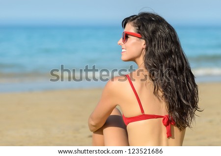 Beautiful brunette young woman sitting on the beach and watching the blue sea. Caucasian beautiful model wearing red bikini and sunglasses. Copy space.