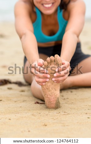 Female hands grabbing up foot sole and stretching leg. Unrecognizable young woman exercising on beach.
