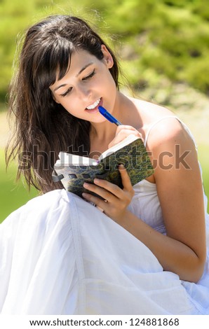 Young brunette woman sitting in nature for reading and writing in her diary. Caucasian model looking interested.