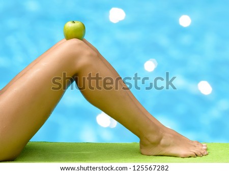 Beautiful female legs with an apple on knees on blue water background. Diet summer concept.