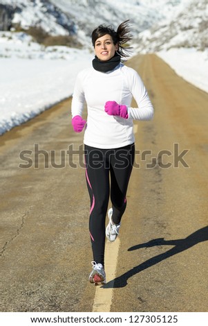 Winter running woman on mountain road. Caucasian fitness girl athlete with warm sport clothes exercising in snowy landscape.