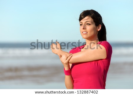 Fitness woman warming up and stretching arm and shoulder on the beach. Female athlete working out outdoors. Hispanic caucasian model.