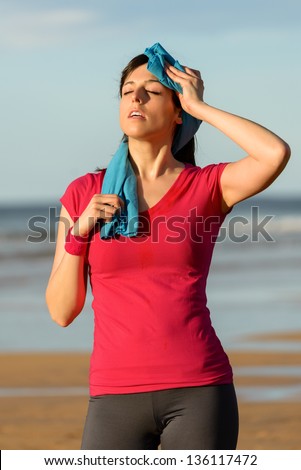 Athlete woman wiping sweat from her forehead with a towel after running in summer on beach . Tired fitness girl sweating after exercising outdoors.