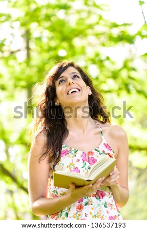 Happy woman reading and holding an open story book in fresh green park on spring or summer day. Caucasian woman smiling and enjoying outdoors.