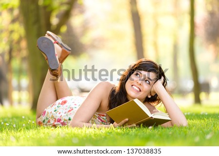 Happy woman reading and holding  story book in fresh green park on spring or summer. Caucasian brunette beautiful girl smiling and day dreaming lying down outdoors.