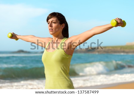 Woman training shoulders with dumbbells on beach. Summer work out, fitness and exercising with weights outdoor. Caucasian sport girl training hard.