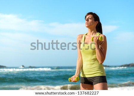Fitness woman working out on beach in summer. Sporty girl training biceps hard with dumbbells. Sweaty sport caucasian female sportswoman.