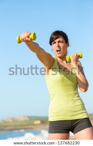 Fitness woman working out on beach in summer. Sporty girl punching and boxing with dumbbells. Hard exercising and training.