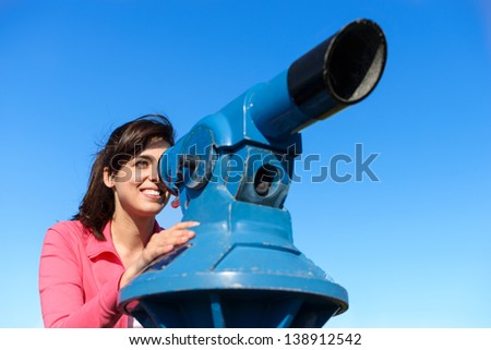 Happy woman looking through telescope on clear summer blue sky background. Beautiful and cheerful caucasian tourist enjoying her travel watching around.