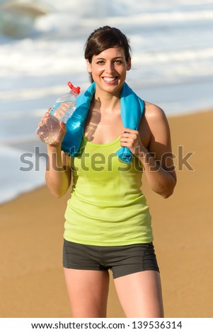 Happy fitness beautiful woman drinking water and sweating after exercising on summer hot day in beach. Female athlete smiling after work out on beach.