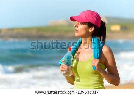 Fitness beautiful woman holding water bottle and sweating after exercising on summer hot day in beach. Female athlete after work out.