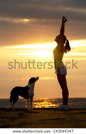 Successful happy woman with her dog raising arms celebrating victory on beautiful golden summer sunset in the beach.