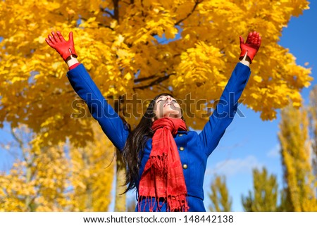Blissful excited woman raising arms to golden yellow trees and praising on autumn day in park or forest. Caucasian fun girl happiness on fall season.