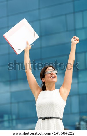 Happy business woman raising arms for celebrating job success on corporate building background. Successful caucasian hispanic corporation worker portrait.