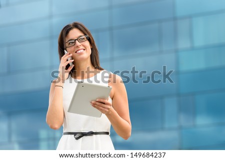 Business woman holding tablet pc computer and calling on cell phone outside corporate building. Caucasian hispanic female executive doing her job using digital communication technology.