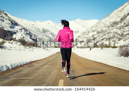 Woman running in cross country road on winter. Rear view of female runner training for marathon in snowy mountains landscape.