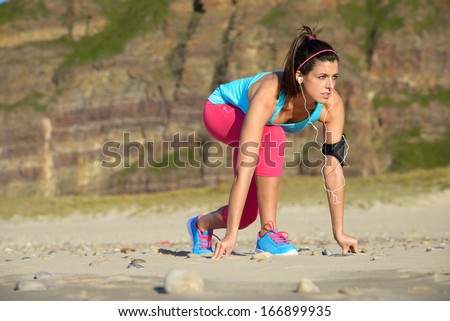 Fitness woman ready for running on beach. Female runner with earphones and arm sport band in starting line pose.