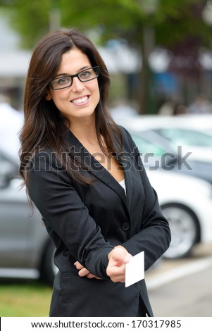 Successful Female Car Sales Representative Giving Blank Business Card In Luxury Automobile Trade Fair. Beautiful Brunette Saleswoman Outdoor.