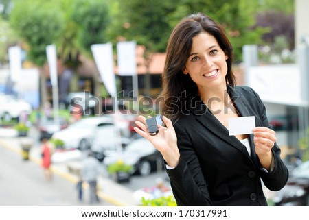 Successful female car sales representative showing car key and business card  in automobile trade fair. Beautiful brunette saleswoman outdoor.