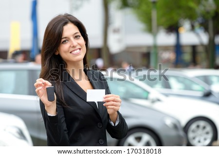 Successful Female Luxury Car Sales Representative Showing Car Key And Business Card In Automobile Trade Fair. Beautiful Brunette Saleswoman Outdoor.