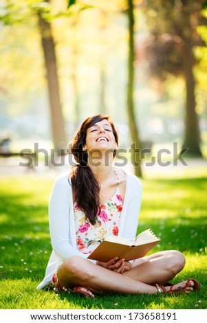 Joyful woman reading fun book sitting on grass in park.