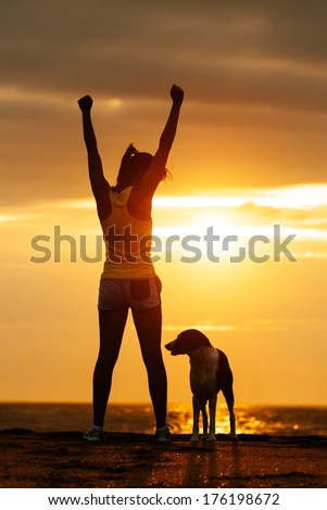 Successful woman and dog enjoying freedom on beautiful golden sunset. Fitness girl raising arms celebrating sport achievement with her pet.