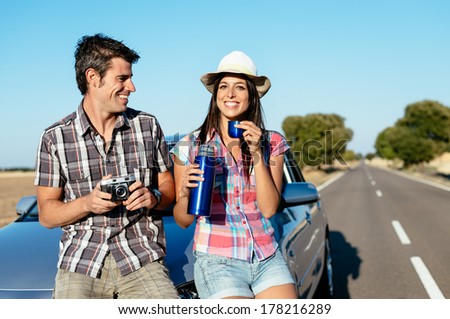 Happy couple on car roadtrip tourism. Man and woman on road travel vacation taking a break for drinking and taking photos.