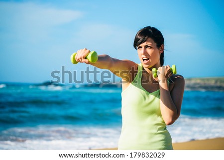 Fitness woman punching hard for working out with dumbbells on beach. Summer intense workout.