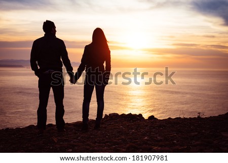 Young couple of lovers holding hands towards the sea at sunset. Man and woman in love looking the sun and ocean together.