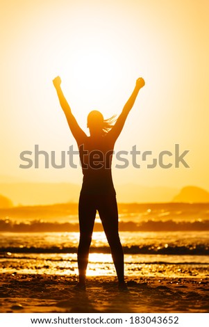 Fitness woman celebrating sport success on beautiful summer sunset or morning on the beach.  Successful female runner silhouette raising arms to the sun.