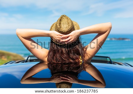 Rear view of relaxed woman on summer travel vacation to the coast  leaning out car sunroof towards the sea.