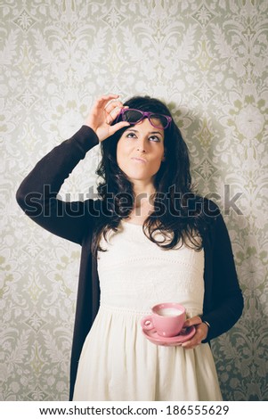 Pensive and suspecting woman leaning on retro ornamental decorated wall.  Brunette girl pulling glasses up and golding a pink cup of coffee. Instagram retro filtered image.