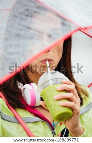 Fitness woman drinking vegetable detox smoothie after workout. Sportswoman sipping healthy drink with straw on rainy day.