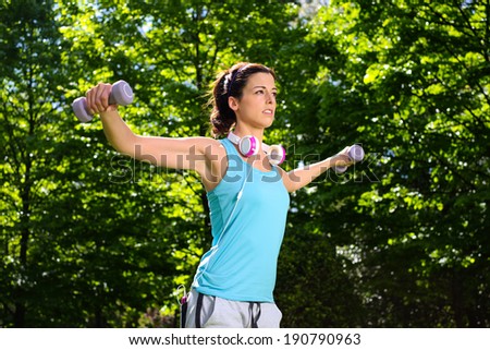 Fitness woman working out shoulder muscles with dumbbells in park. Spring or summer exercising workout with weights. Outdoor sport lifestyle concept.