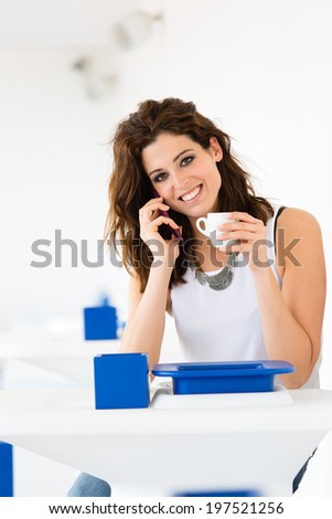 Cheerful relaxed woman talking on cellphone and drinking a cup of coffee in a summer chillout bar. Hispanic brunette casual model.