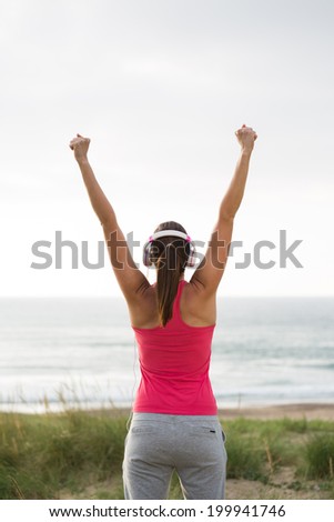 Successful female athlete with headphones raising arms towards the sea. Fitness woman celebrating exercising achievement after workout.