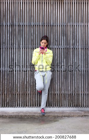 Relaxed urban fitness woman reading or texting with smartphone and leaning on a wooden wall before workout. Female sporty athlete looking her cellphone.