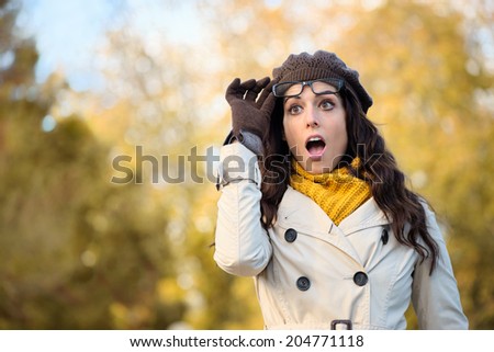 Amazed fashionable woman with mouth open looking surprised raising her glasses in autumn. Eye wear and fall season fashion concept. Beautiful female wearing raincoat, scarf, wool cap and gloves.