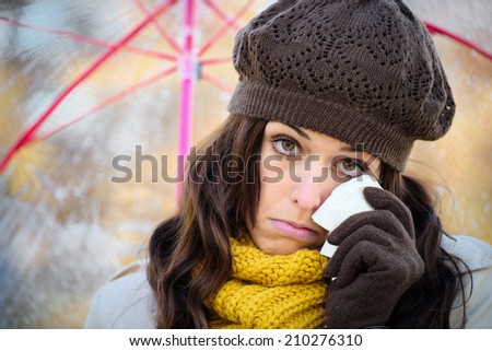 Sad woman in autumn cold rainy day wiping away a tear with a tissue. Depressed female crying and holding umbrella under the rain.