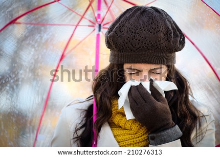 Woman with cold or flu coughing and blowing her nose with a tissue under autumn rain. Brunette female sneezing and wearing warm clothes.