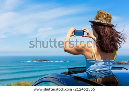 Happy woman taking photos to the sea with smartphone camera on car summer travel vacation to the coast. Brunette girl  leaning out vehicle sunroof towards the ocean.