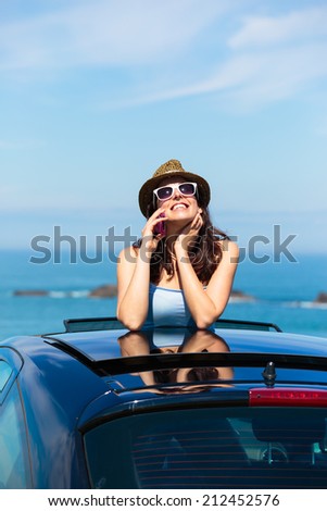 Happy woman on car summer travel calling on cellphone while leaning out sunroof towards the sea. Brunette girl enjoying vacation to the coast.
