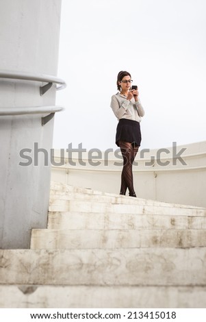 Trendy young businesswoman on a job break in modern corporate building stairs on a working day messaging on smartphone. Brunette professional woman wearing sexy skirt and stockings.